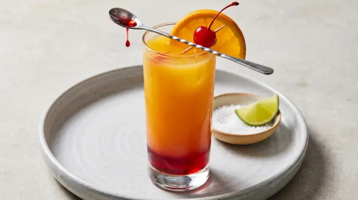 A vibrant red and orange layered Tequila Sunrise cocktail with an orange slice and cherry garnish.