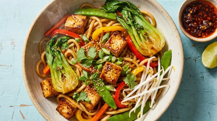 Bowl of vegetarian chow mein with tofu, bok choy, peppers and bean sprouts, chili oil on the side